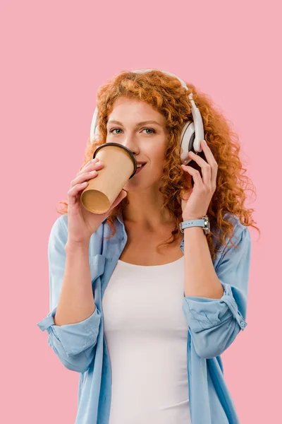 Щаслива руда дівчина п'є каву і слухає музику в навушниках, ізольована на рожевому — стокове фото