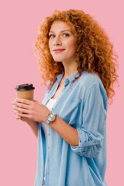 Felice donna riccia con i capelli rossi che tengono il caffè per andare, isolato su rosa — Foto stock