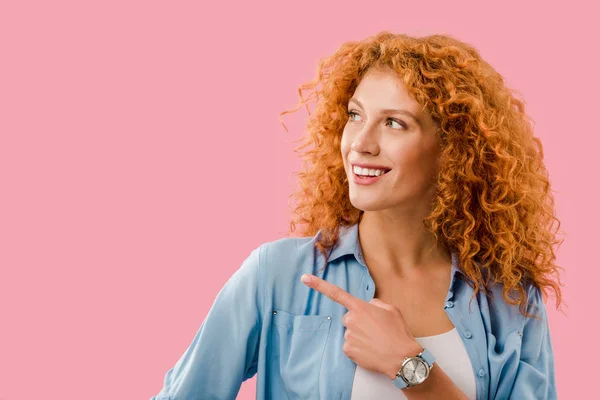 Attractive cheerful redhead woman pointing at something isolated on pink — Stock Photo