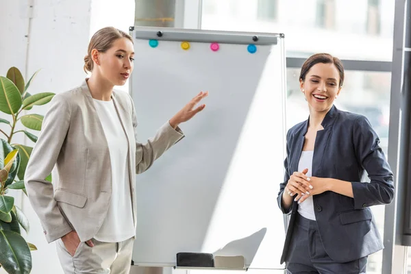 Dos mujeres de negocios sonrientes en ropa formal de pie cerca de flipchart en la oficina - foto de stock