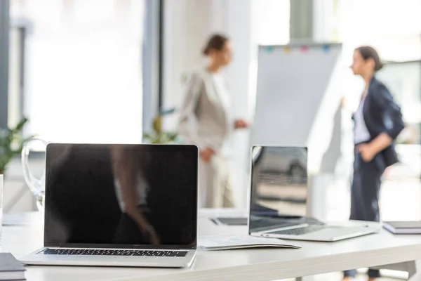 Messa a fuoco selettiva di due donne d'affari in piedi vicino flipchart e laptop sul tavolo in primo piano in ufficio — Foto stock