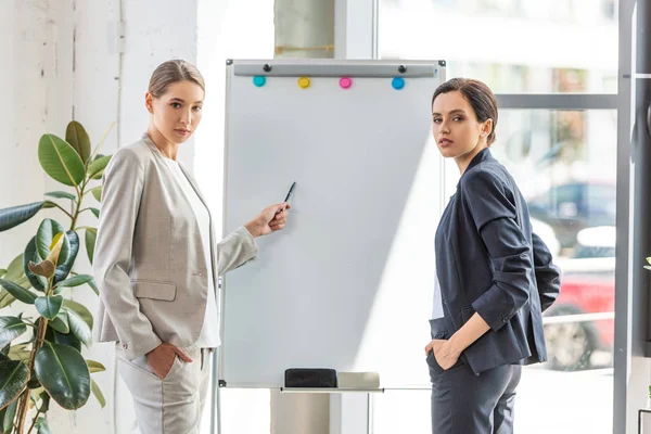 Two businesswomen in formal wear standing near flipchart in office — Stock Photo
