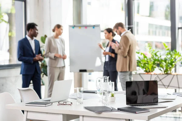 Foco seletivo de quatro empresários multiétnicos perto de flipchart e mesa com laptops e copos de água em primeiro plano — Fotografia de Stock