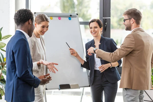 Four smiling multiethnic colleagues in formal wear standing near flipchart in office — Stock Photo