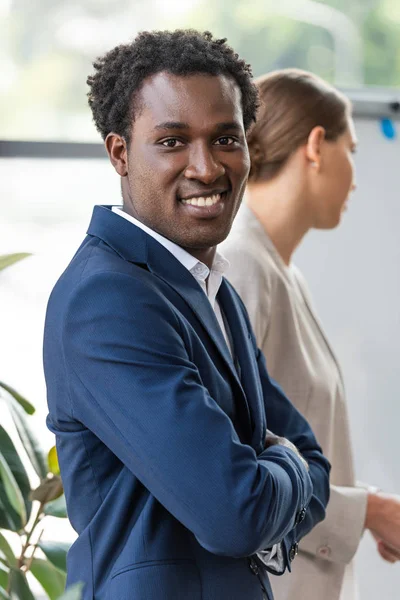 Sonriente hombre de negocios afroamericano de pie con los brazos cruzados cerca de colega y mirando a la cámara - foto de stock