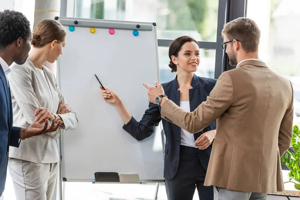 Four smiling multiethnic colleagues in formal wear standing near flipchart in office — Stock Photo