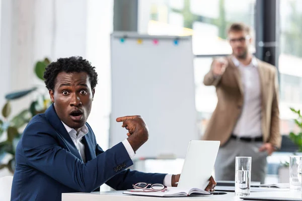 Schockierter afrikanisch-amerikanischer Geschäftsmann sitzt am Tisch und zeigt im Büro mit dem Finger auf sich selbst — Stockfoto