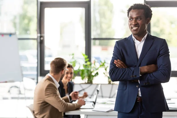 Sonriente hombre de negocios afroamericano en ropa formal de pie con los brazos cruzados delante de sus colegas en la oficina - foto de stock
