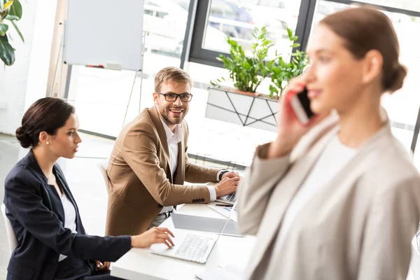 Enfoque selectivo de la mujer de negocios de pie cerca de colegas y hablando en el teléfono inteligente en la oficina - foto de stock