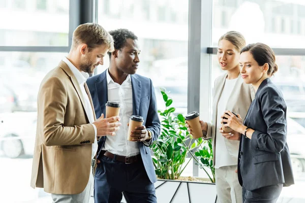 Quatre collègues multiethniques tenant des tasses jetables de café et souriant au bureau — Photo de stock