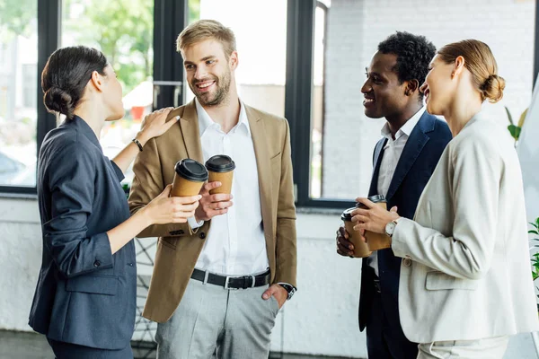 Quatre collègues multiethniques tenant des tasses jetables de café et souriant au bureau — Photo de stock