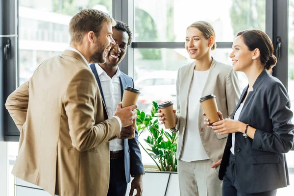 Cuatro colegas multiétnicos sosteniendo tazas de café desechables y sonriendo en la oficina - foto de stock