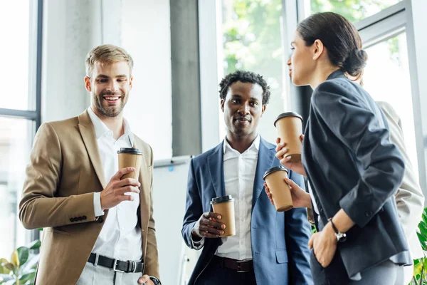 Three multiethnic colleagues holding disposable cups of coffee and smiling in office — Stock Photo