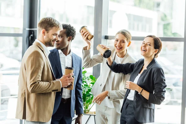 Four multiethnic colleagues holding disposable cups of coffee and smiling in office — Stock Photo