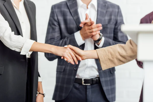 Vista recortada de tres colegas en ropa formal estrechando la mano en la sala de conferencias - foto de stock