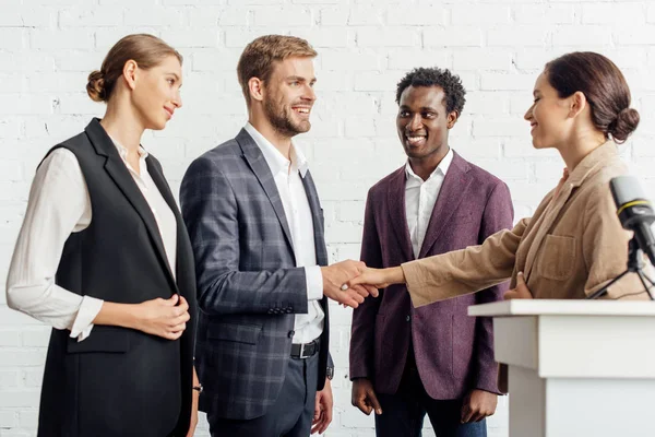 Four multiethnic colleagues in formal wear talking and shaking hands in conference hall — Stock Photo