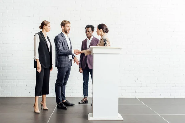 Cuatro colegas multiétnicos en ropa formal hablando y dándose la mano en la sala de conferencias - foto de stock