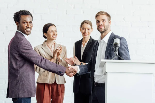 Four multiethnic colleagues in formal wear talking and shaking hands in conference hall — Stock Photo