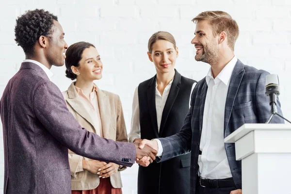 Cuatro colegas multiétnicos en ropa formal hablando y dándose la mano en la sala de conferencias - foto de stock