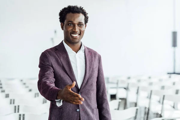 Hombre de negocios afroamericano en ropa formal con la mano extendida sonriendo en la sala de conferencias - foto de stock