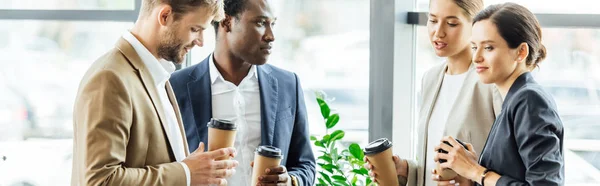 Plano panorámico de cuatro colegas multiétnicos sosteniendo tazas de café desechables y hablando en la oficina - foto de stock