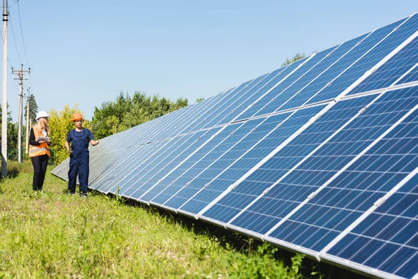 Ingeniero guapo y atractiva mujer de negocios hablando y caminando cerca de baterías de energía solar - foto de stock