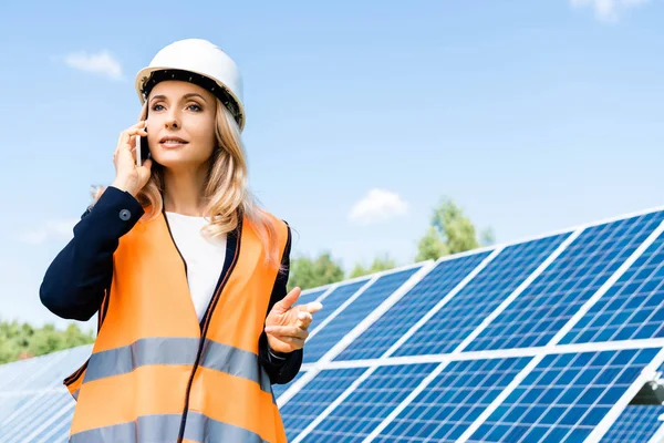 Atractiva mujer de negocios en hardhat y chaleco de seguridad hablando en el teléfono inteligente - foto de stock