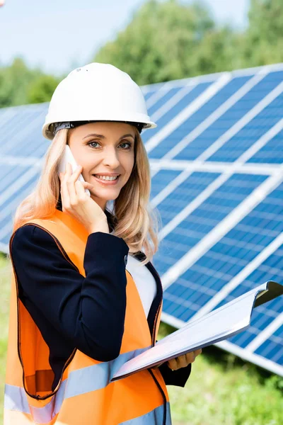 Atractiva mujer de negocios en hardhat y chaleco de seguridad hablando en el teléfono inteligente — Stock Photo