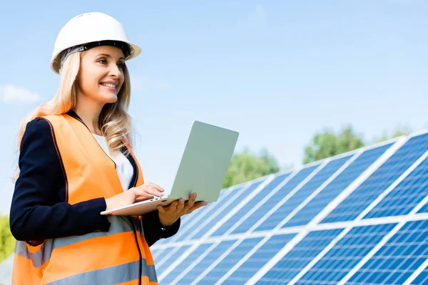 Mujer de negocios en chaleco de seguridad y hardhat sonriendo y utilizando el ordenador portátil - foto de stock