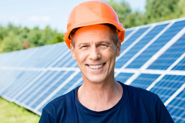 Bel ingénieur en t-shirt et casque orange souriant et regardant la caméra — Photo de stock