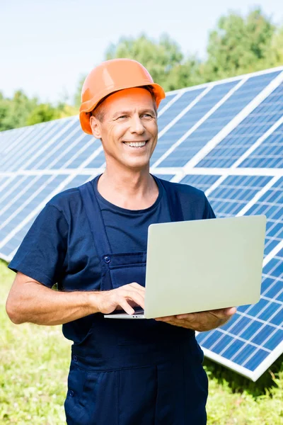 Handsome engineer in t-shirt and orange hardhat smiling and holding laptop — Stock Photo