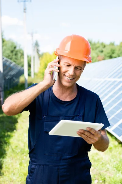 Handsome engineer in t-shirt and orange hardhat smiling and talking on smartphone — Stock Photo