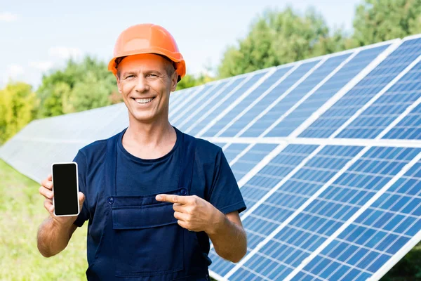 Ingeniero guapo en camiseta y naranja hardhat sonriendo y señalando con el dedo en el teléfono inteligente - foto de stock