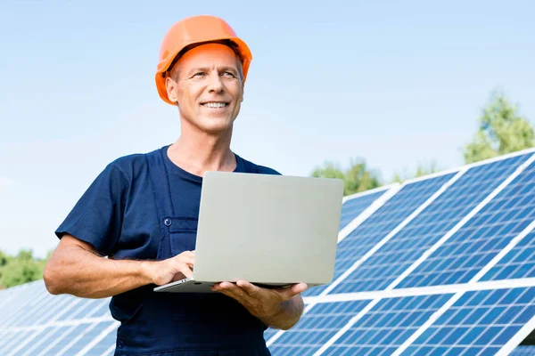 Ingeniero guapo en camiseta y naranja hardhat sonriendo y sosteniendo portátil - foto de stock