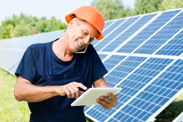 Ingeniero guapo en camiseta y naranja hardhat sonriendo y hablando en el teléfono inteligente - foto de stock