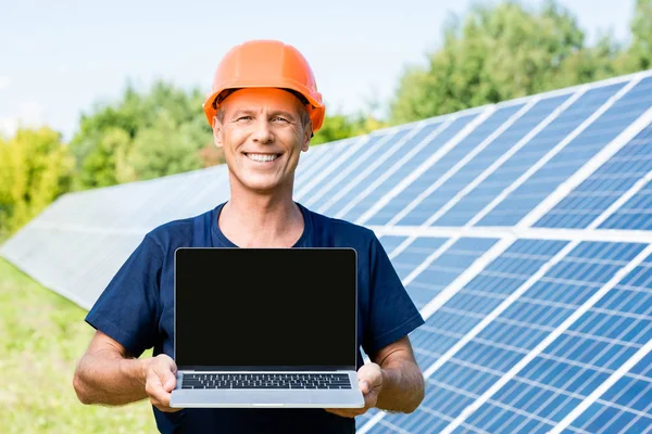 Engenheiro bonito em t-shirt e hardhat laranja sorrindo e segurando laptop — Fotografia de Stock