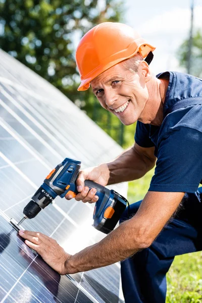 Handsome engineer in t-shirt and orange hardhat smiling and using drill — Stock Photo