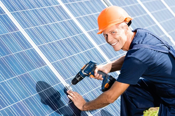 Ingeniero guapo en camiseta y naranja hardhat sonriendo y utilizando taladro - foto de stock