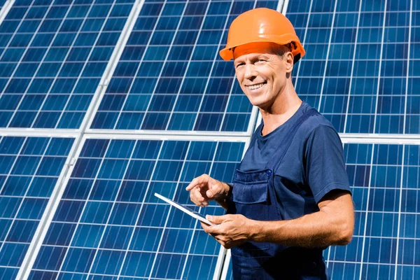 Handsome engineer in t-shirt and orange hardhat smiling and holding digital tablet — Stock Photo