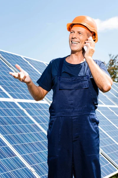 Ingeniero guapo en camiseta y naranja hardhat sonriendo y hablando en el teléfono inteligente - foto de stock