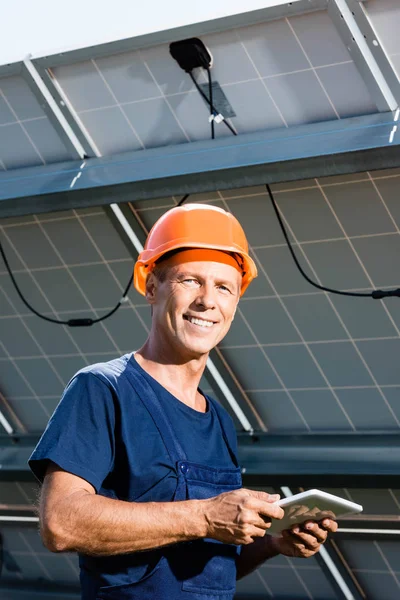 Handsome engineer in t-shirt and orange hardhat smiling and holding digital tablet — Stock Photo