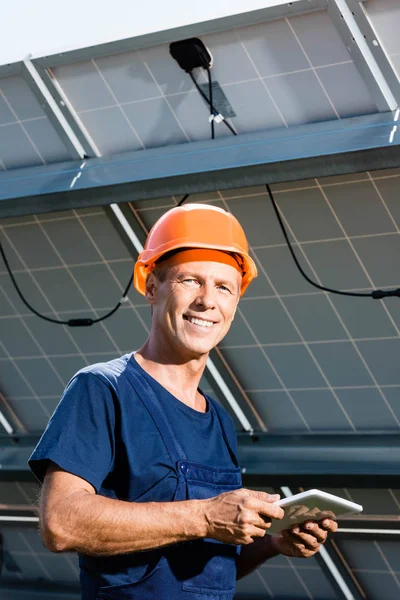 Schöner Ingenieur in T-Shirt und orangefarbener Mütze, lächelnd und in der Hand ein digitales Tablet — Stockfoto