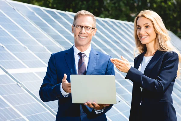 Guapo hombre de negocios y atractiva mujer de negocios sonriendo y sosteniendo portátil - foto de stock