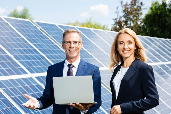 Guapo hombre de negocios y atractiva mujer de negocios sonriendo y sosteniendo portátil - foto de stock