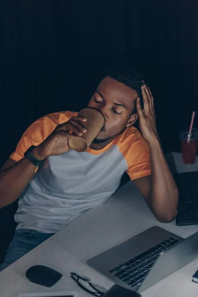 Young african american programmer drinking coffee to go while working at night in office — Stock Photo