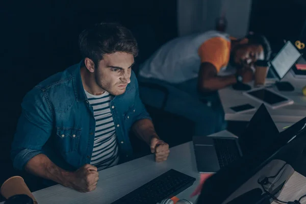 Programmatore irritato gesticolare mentre lavora di notte in ufficio vicino a collega afro-americano che dorme sul posto di lavoro — Foto stock