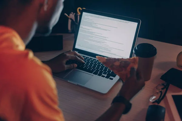 Vista recortada del joven programador afroamericano comiendo pizza mientras trabaja de noche en la oficina - foto de stock