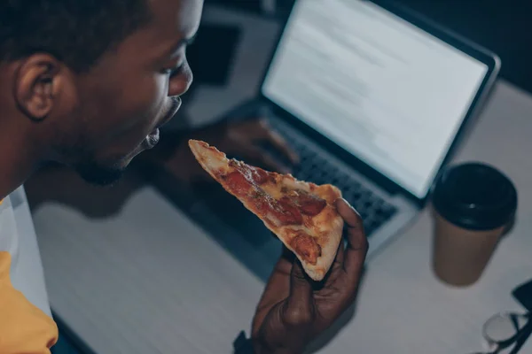 Jovem programador afro-americano comendo pizza enquanto trabalhava à noite no escritório — Fotografia de Stock