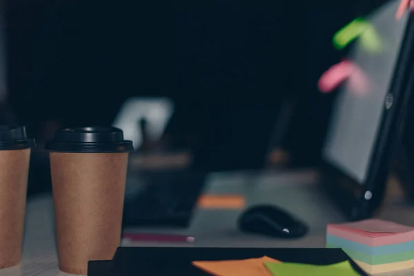 Escritorio de oficina con café para llevar, monitor de computadora, ratón de computadora y cuadernos por la noche - foto de stock