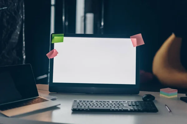 Escritorio de oficina con monitor de ordenador, ordenador portátil, teclado y notas adhesivas por la noche - foto de stock